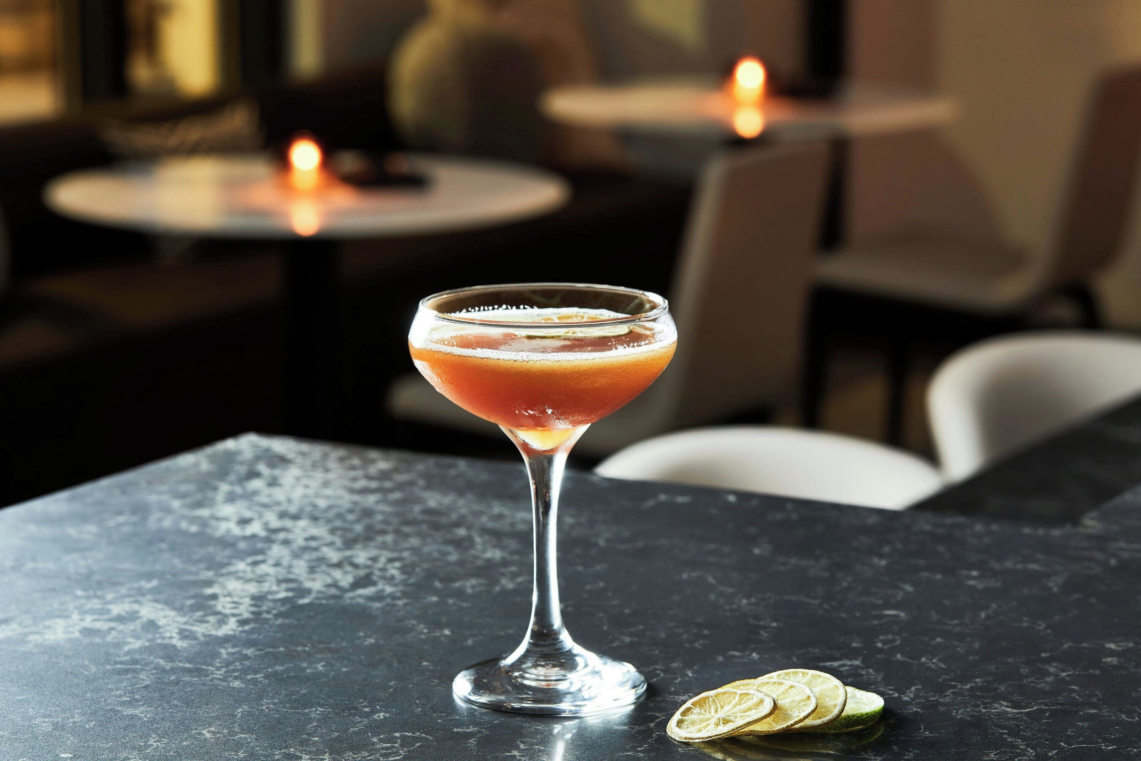 A cocktail in a martini glass on a dark marble table with a slice of lime and a lemon wedge beside it.