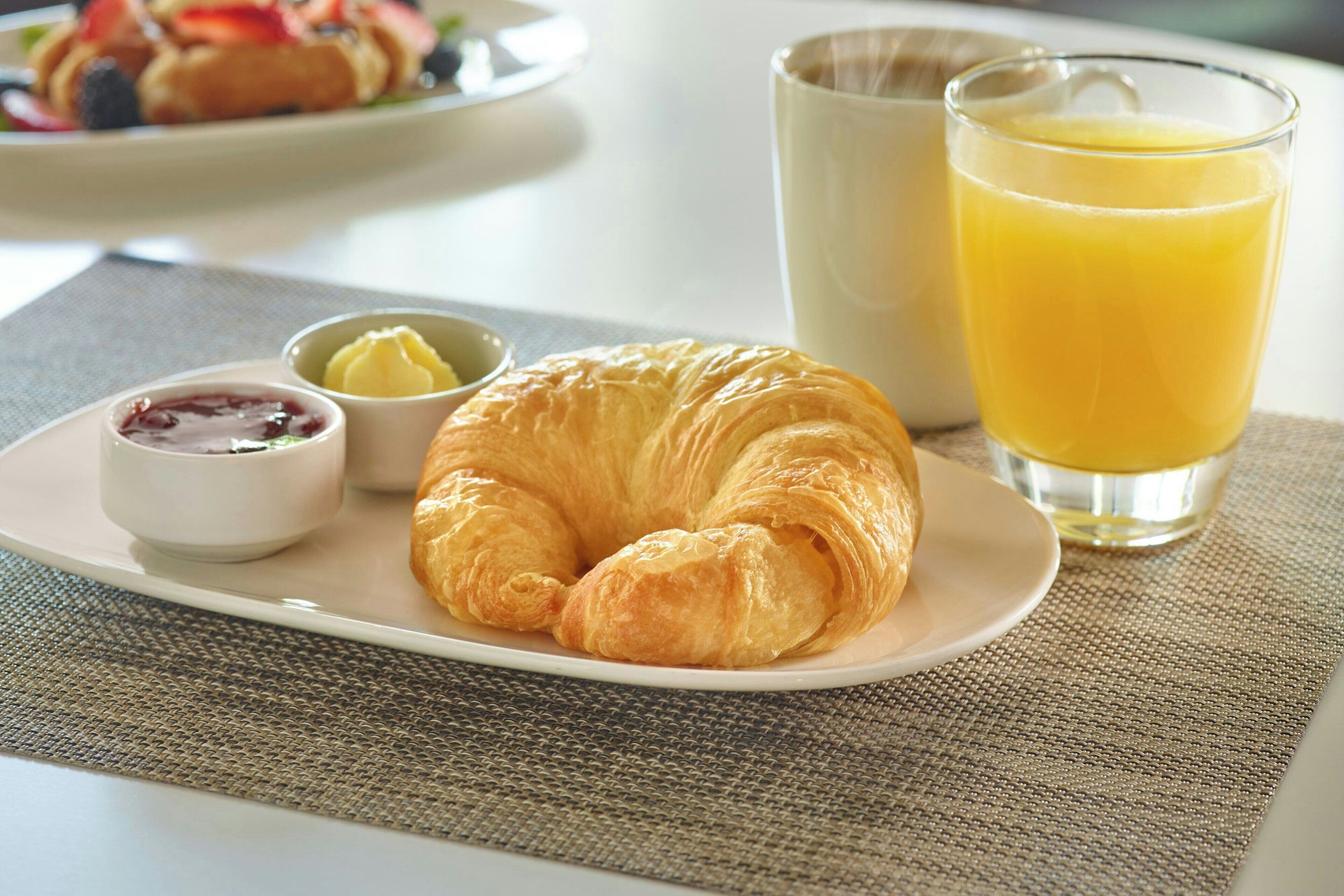 Une table dressée pour le petit-déjeuner, avec un croissant, du beurre, de la confiture et du jus d’orange sur une assiette.