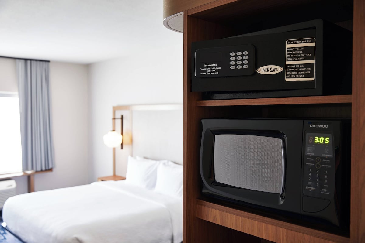 A hotel room with a bed, a wooden shelf with a microwave and a control panel, and a window with curtains.