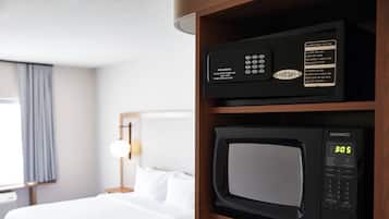 A hotel room with a bed, a wooden shelf with a microwave and a control panel, and a window with curtains.