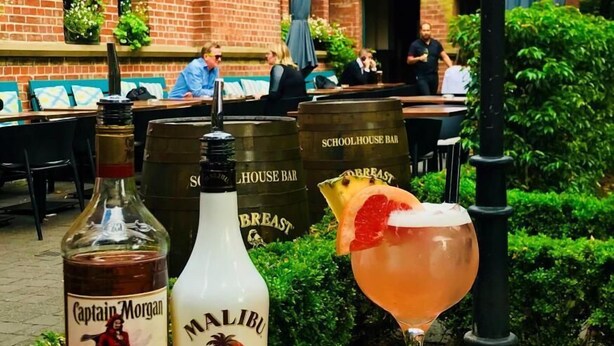 Uma mesa com uma garrafa de Captain Morgan, uma garrafa de Malibu e um cocktail com decoração de grapefruit, em frente a um edifício de tijolos com uma placa indicando um restaurante.