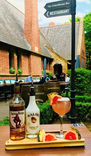 A table with a bottle of Captain Morgan, a bottle of Malibu, and a cocktail with a grapefruit garnish, set against a brick building with a sign pointing to a restaurant. - Schoolhouse Hotel (Dublin)