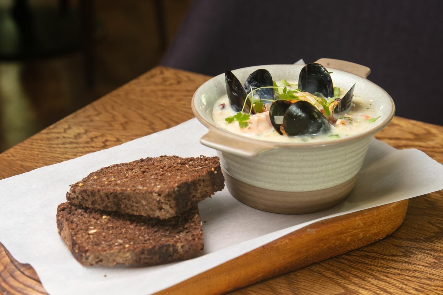 Un plato de mejillones con salsa cremosa y una rebanada de pan de centeno oscuro sobre una mesa de madera.