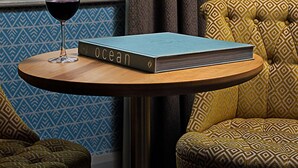 Quarto com vista para árvores e edifícios pela janela, uma mesa de madeira redonda com um livro intitulado 'ocean' e um copo de vinho tinto, e mobiliário estampado.