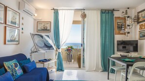 Un salón con un sofá azul, una mesa de comedor con sillas, un televisor y vista al mar a través de un balcón.