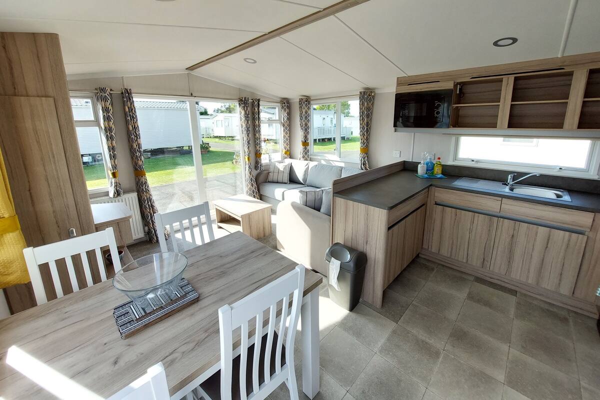 A modern kitchen with a microwave, sink, and wooden cabinets. There is a dining area with a table and chairs, and a view of a grassy yard through large windows.
