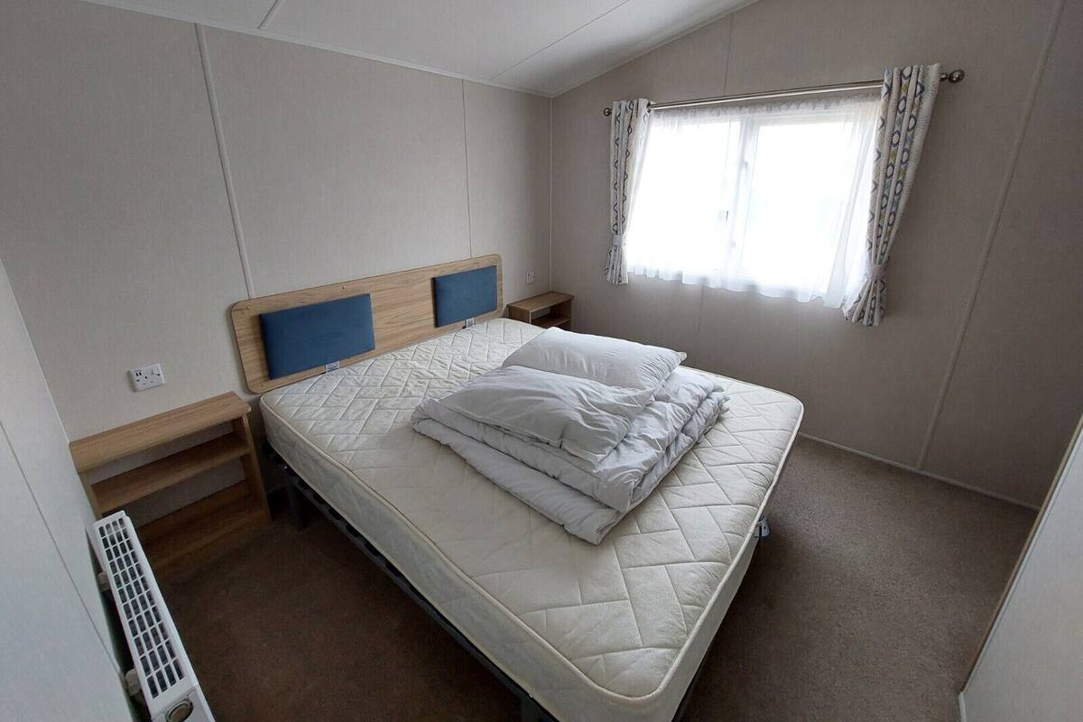 A single bed with a quilted mattress, a headboard with blue panels, a bedside table, a power outlet, a window with curtains, and a radiator.