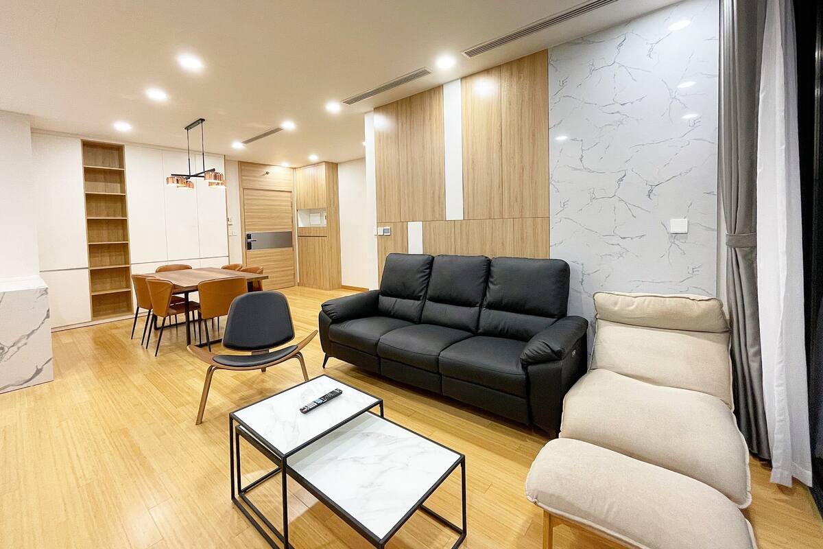 A modern living room with a black leather sofa, a beige armchair, a small white table, and a dining area with a wooden table and chairs.