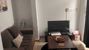 A living room with a brown sofa, a TV on a stand, a fan, and a table with a bowl and papers. - BCBG KinShasa Apartment (Limete)