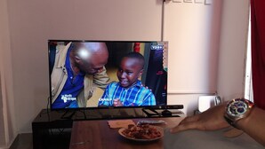 A television displaying a news program, a wooden table with a bowl of food, and a remote control. - BCBG KinShasa Apartment (Limete)
