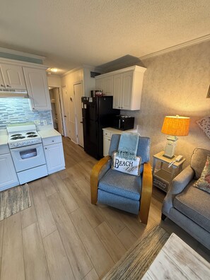 A compact living space with a kitchenette, a blue armchair with a beach-themed pillow, a lamp, and a sofa.