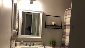 Un bany amb un lavabo blanc, un mirall, una petita estant amb productes de tocador, un vàter i una cortina de bany.