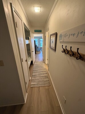 A hallway with a wooden floor, a rug, a door leading to a room, and a wall with a sign and hooks.
