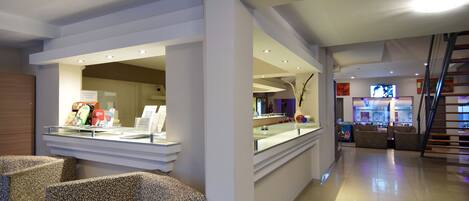 A hotel lobby with a reception desk, patterned seating, a staircase, and a kitchen area in the background.
