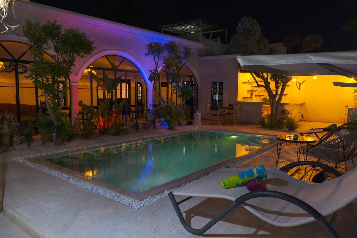 A pool area with lounge chairs and umbrellas, surrounded by a building with arched windows and a covered patio.