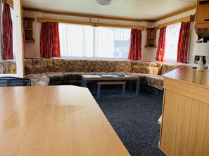 A cozy RV interior with a sofa set, a wooden table, and a kitchen area with a lamp. - Fantasy Island Caravan Hire (Skegness)