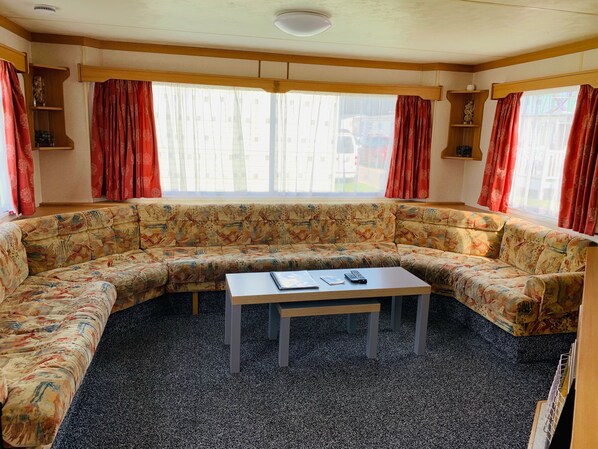 A cozy living room with a large floral-patterned sofa, a small wooden coffee table, and red curtains. - Fantasy Island Caravan Hire (Skegness)