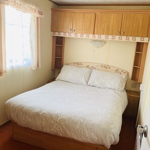 A bedroom with a bed, wooden headboard, built-in shelving, and a window with curtains. - Fantasy Island Caravan Hire (Skegness)