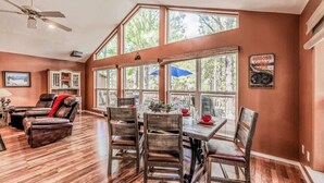 A spacious living room with a dining area, large windows, and a ceiling fan. - Four Bears Bungalow (Ruidoso)