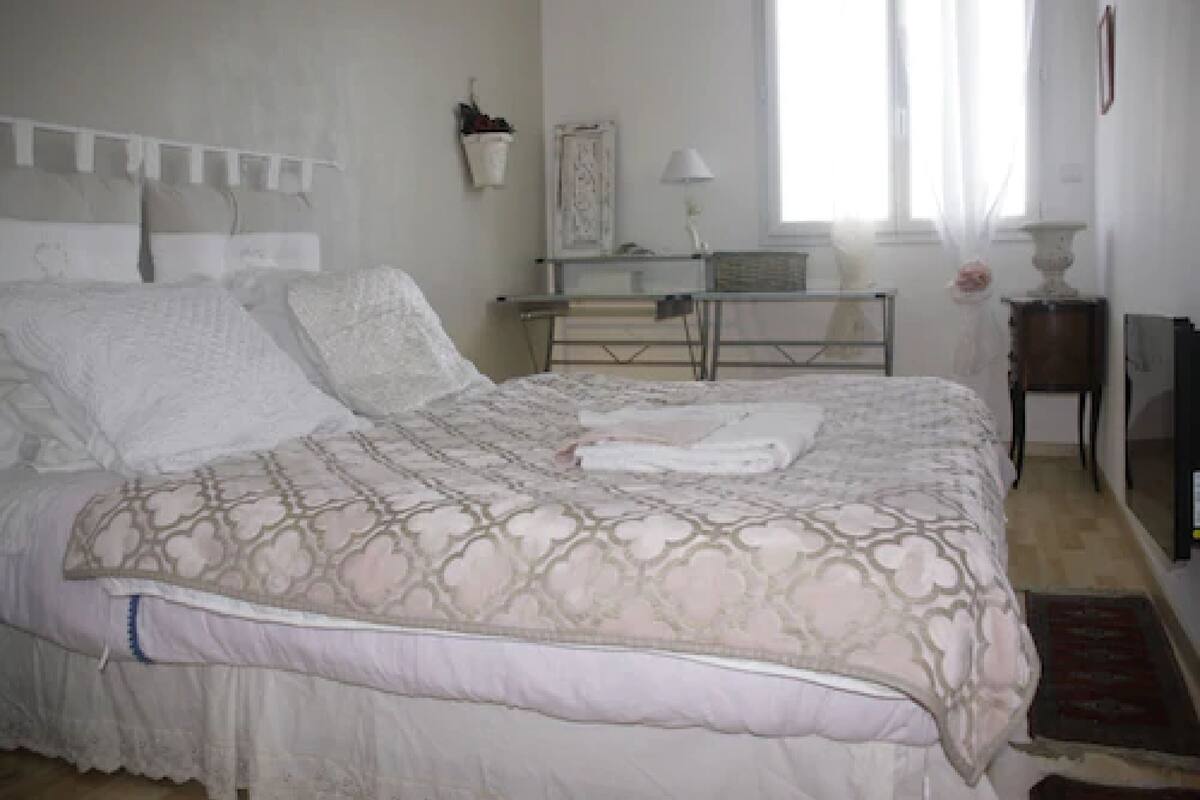 A neatly made bed with a patterned bedspread, a bedside table with a lamp, and a window with sheer curtains.