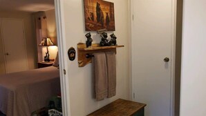 A bedroom with a bed, a nightstand with a lamp, a wooden shelf with decorative items, and a door with a 'Welcome' sign. - Papa Bears Cabin (Ruidoso)
