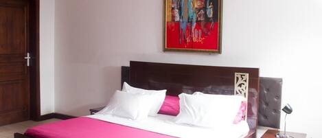 Una habitación de hotel con una cama de madera, ropa de cama blanca y rosa, un cuadro enmarcado en la pared y una mesita de noche con una lámpara.