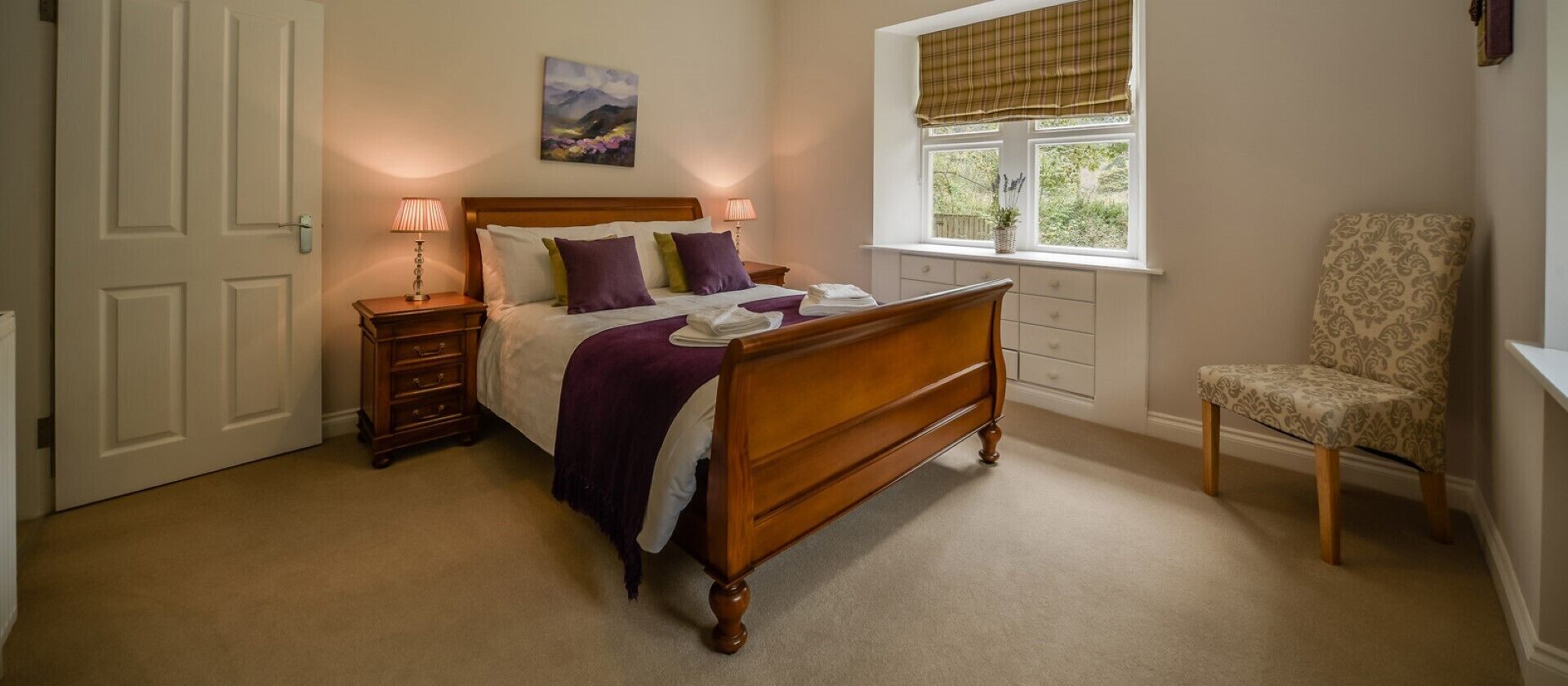 A bedroom with a wooden bed, bedside tables, a chair, a window with a view, and a painting on the wall.