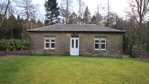 Ein kleines, zweistöckiges Steinhaus mit weißer Tür und Fenstern, umgeben von einem Rasen und Bäumen.