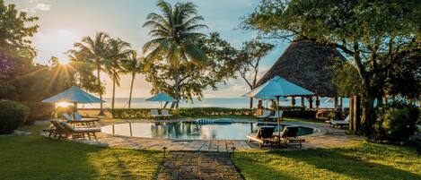 A pool area with lounge chairs and umbrellas, surrounded by palm trees and a view of the ocean.