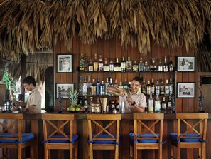 A bar with wooden shelves stocked with liquor bottles, framed pictures on the wall, and blue cushioned bar stools.