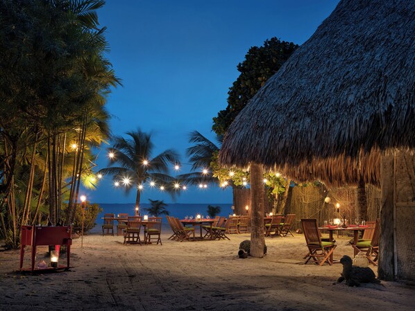 A beachside dining area with tables and chairs, palm trees, and a thatched-roof structure.