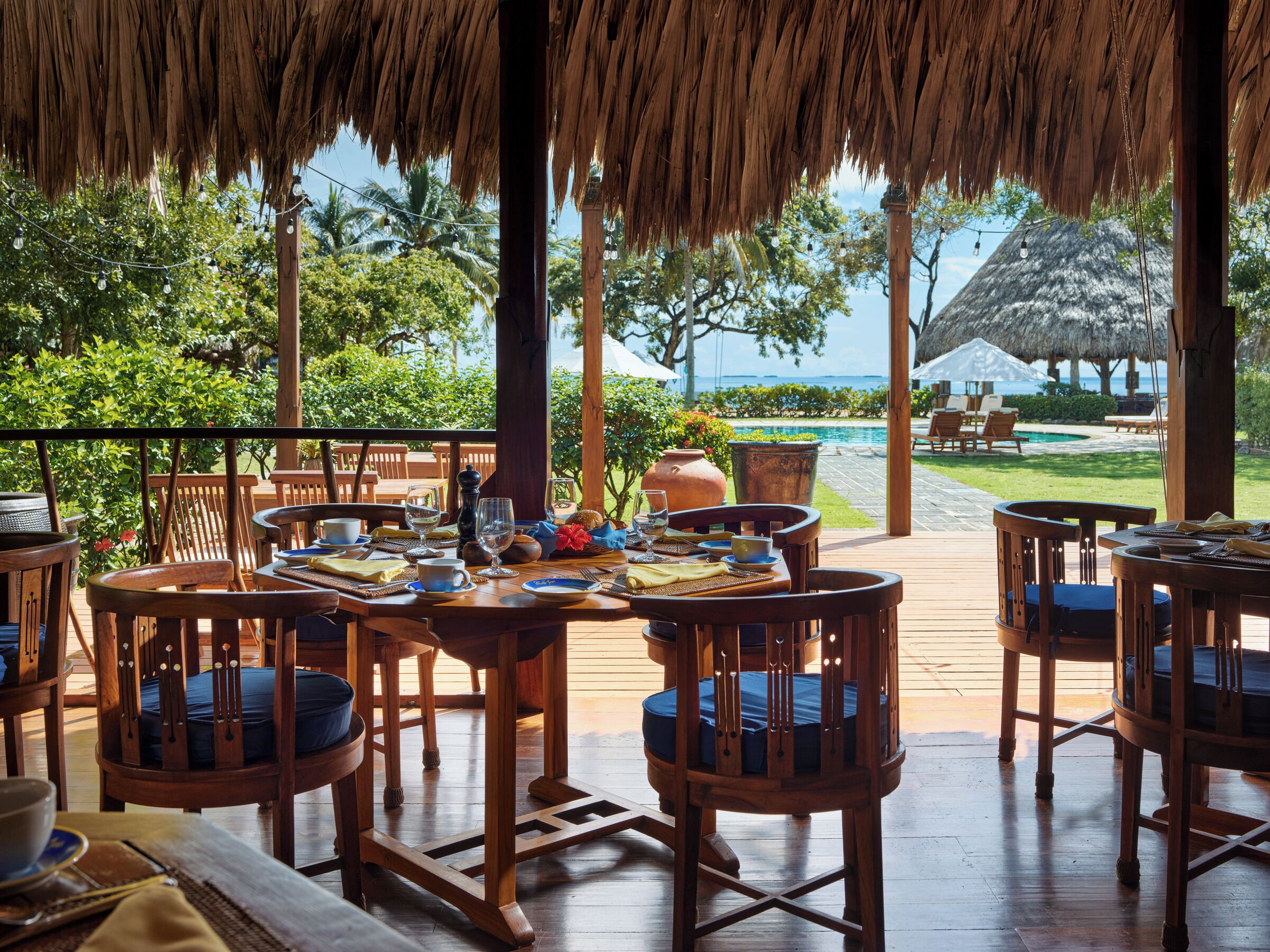 An outdoor dining area with wooden tables and chairs, a thatched roof, and a view of a pool and palm trees.