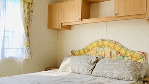 A bedroom with a bed, two nightstands, wooden cabinets above the bed, and a window with curtains. - Golden Sands Caravan Hire (Skegness)