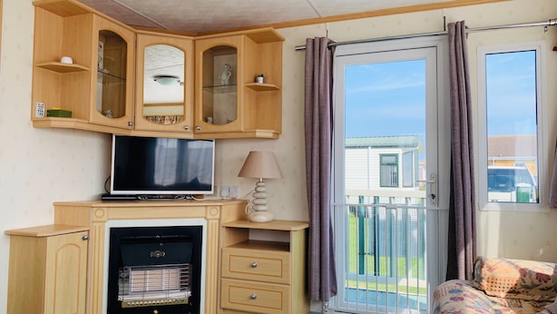 A room with a television, a fireplace, wooden cabinets, a lamp, and a balcony with a view of a house. - Golden Sands Caravan Hire (Skegness)