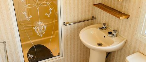 A bathroom with a shower, sink, toilet, and a wooden shelf above the sink.