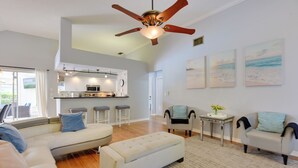A spacious living area with a sofa, ottoman, and armchairs, a kitchen with bar stools, and a ceiling fan.