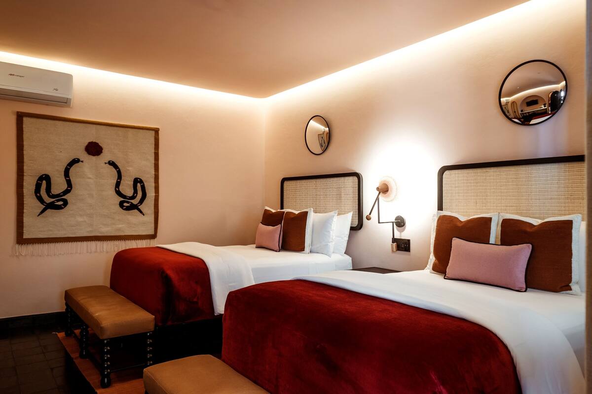 Ein Hotelzimmer mit zwei Betten, jeweils mit roter und weißer Bettwäsche, einem großen abstrakten Kunstwerk an der Wand, einem runden Spiegel und einem kleinen Tisch mit einer Lampe.