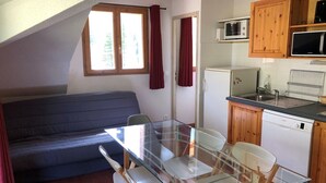 Una cocina compacta con microondas, refrigerador y lavavajillas Bosch. Hay una mesa de comedor de vidrio con sillas, un sofá azul y una ventana con cortinas.