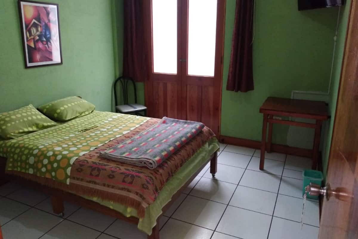 Une chambre à coucher avec un lit, une chaise, une petite table, une porte et une fenêtre avec des rideaux.