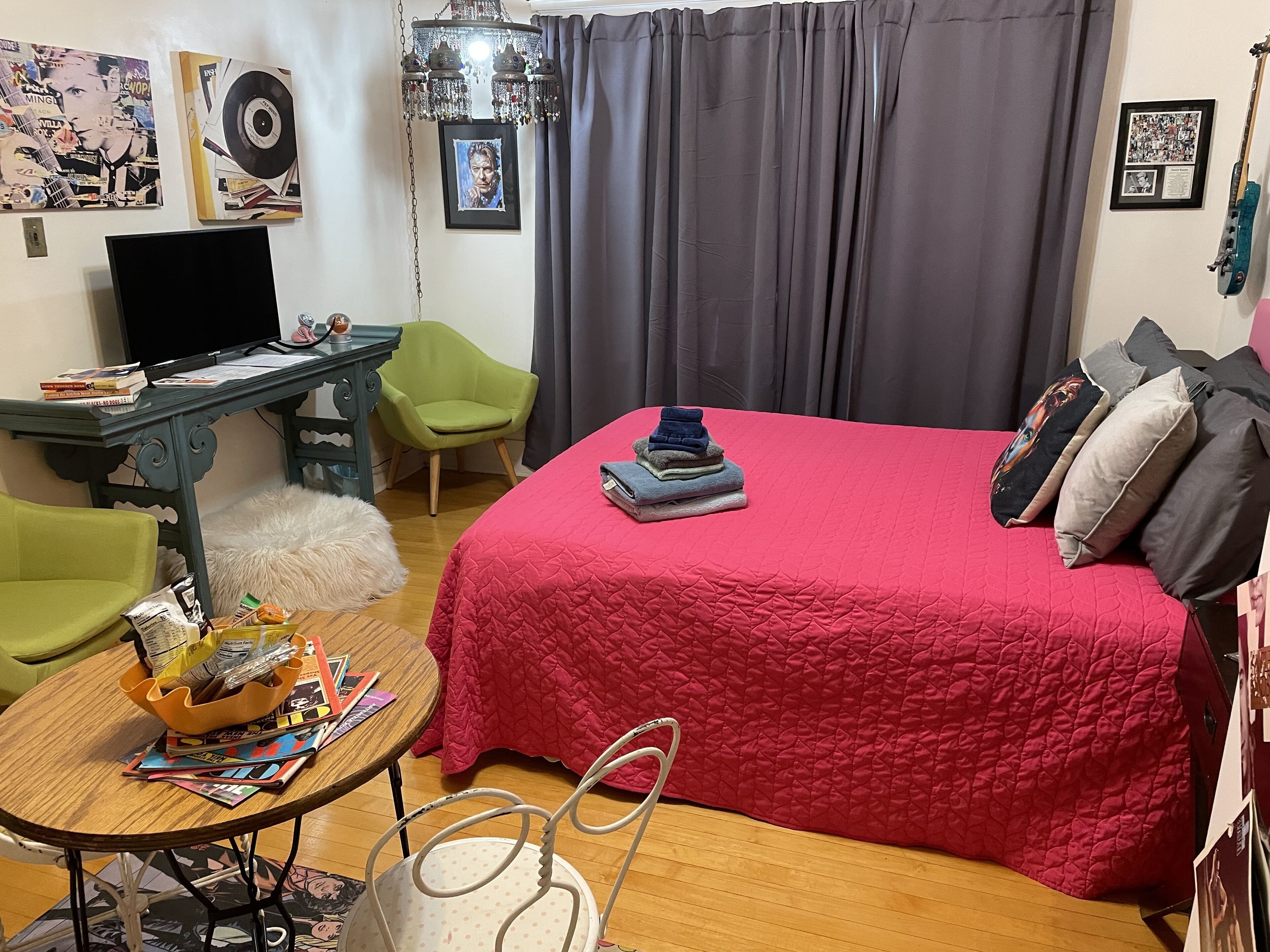 A bedroom with a red bedspread, a green chair, a wooden table with magazines, and a desk with a computer and books.