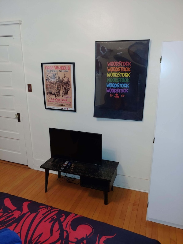 A room with a white door, a framed poster on the wall, a black TV stand with a flat-screen TV, and a patterned rug. - Greenway Manor (Bisbee)