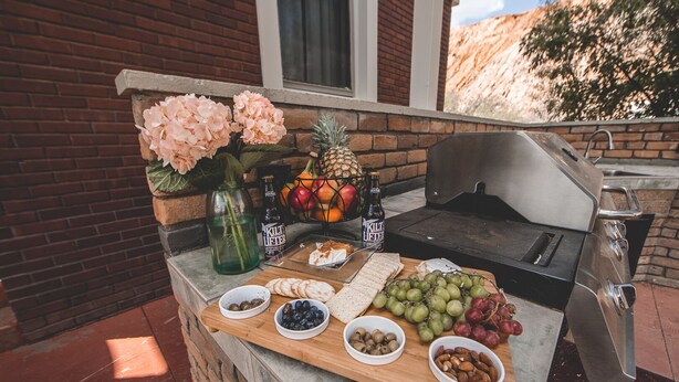 An outdoor barbecue setup with a stone countertop, a grill, and various food items including fruits, cheeses, and nuts. - Greenway Manor (Bisbee)
