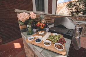 An outdoor barbecue setup with a stone countertop, a grill, and various food items including fruits, cheeses, and nuts. - Greenway Manor (Bisbee)