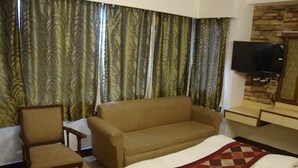 Een hotelkamer met een bank, een stoel, een bed met quilt, een aan de muur bevestigde televisie en gordijnen met een bladpatroon.