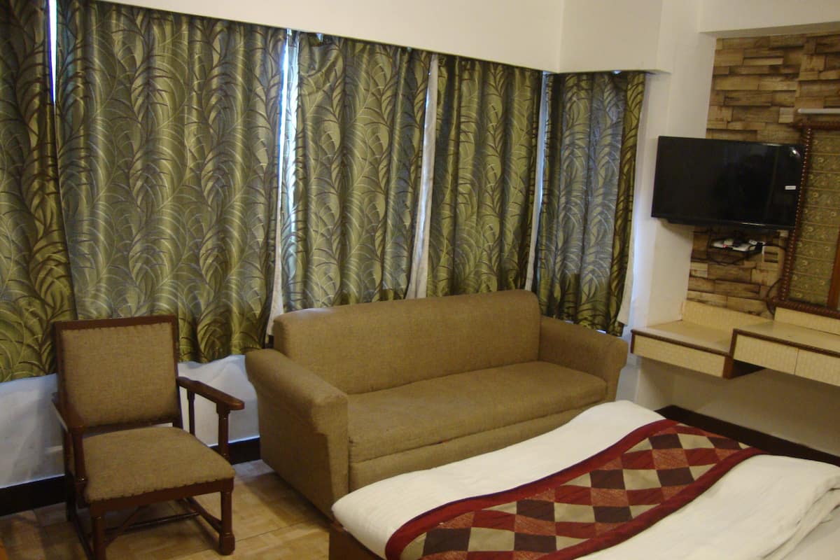 Quarto de hotel com sofá, uma cadeira, cama com colcha e televisão fixada na parede, cortinas com padrão de folhas.