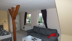Un espacio habitable compacto con un sofá gris, una mesita de centro blanca, una cama con colcha estampada, una silla de madera y una ventana con cortinas.