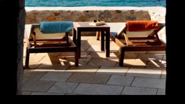 An outdoor seating area with two wooden lounge chairs and a small table, overlooking a clear blue sea and a rocky hill. - Pirgos Mavromichali (East Mani)