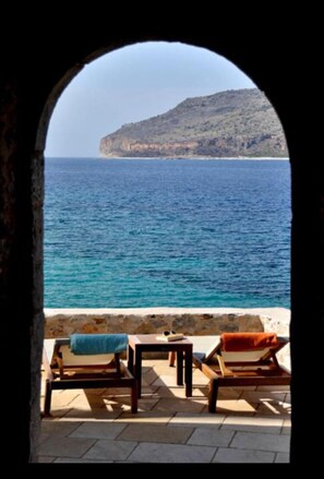 Ein Außenbereich mit zwei Holzliegen und einem kleinen Tisch, mit Blick auf das klare, blaue Meer und einen felsigen Hügel.