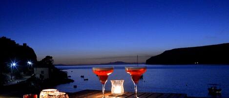 Una mesa con dos vasos de bebida roja y una vela, con vistas a un mar tranquilo al atardecer.