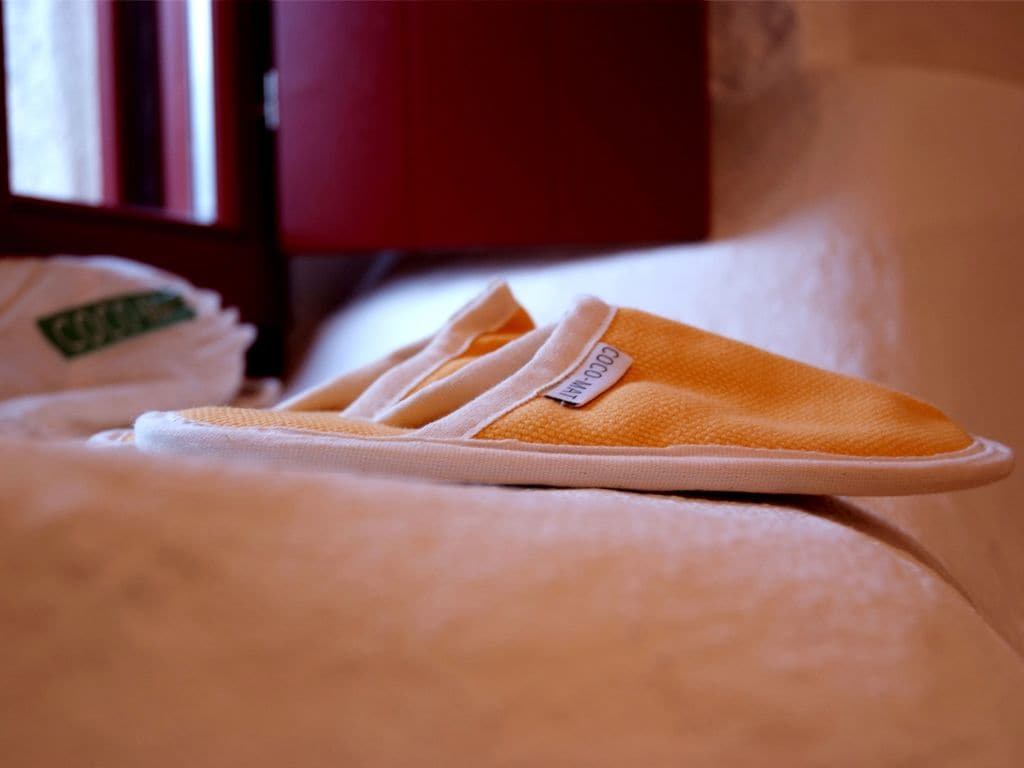 Un par de pantuflas naranjas sobre una cama con un almohadón y una manta blancos.